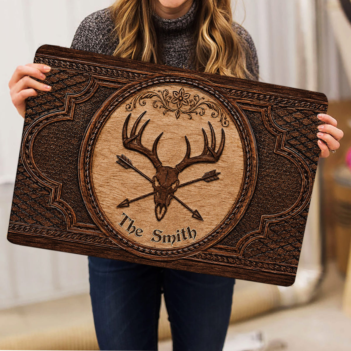 Person holding an ornate hunting doormat with deer skull, antlers, arrows, and rustic brown artwork