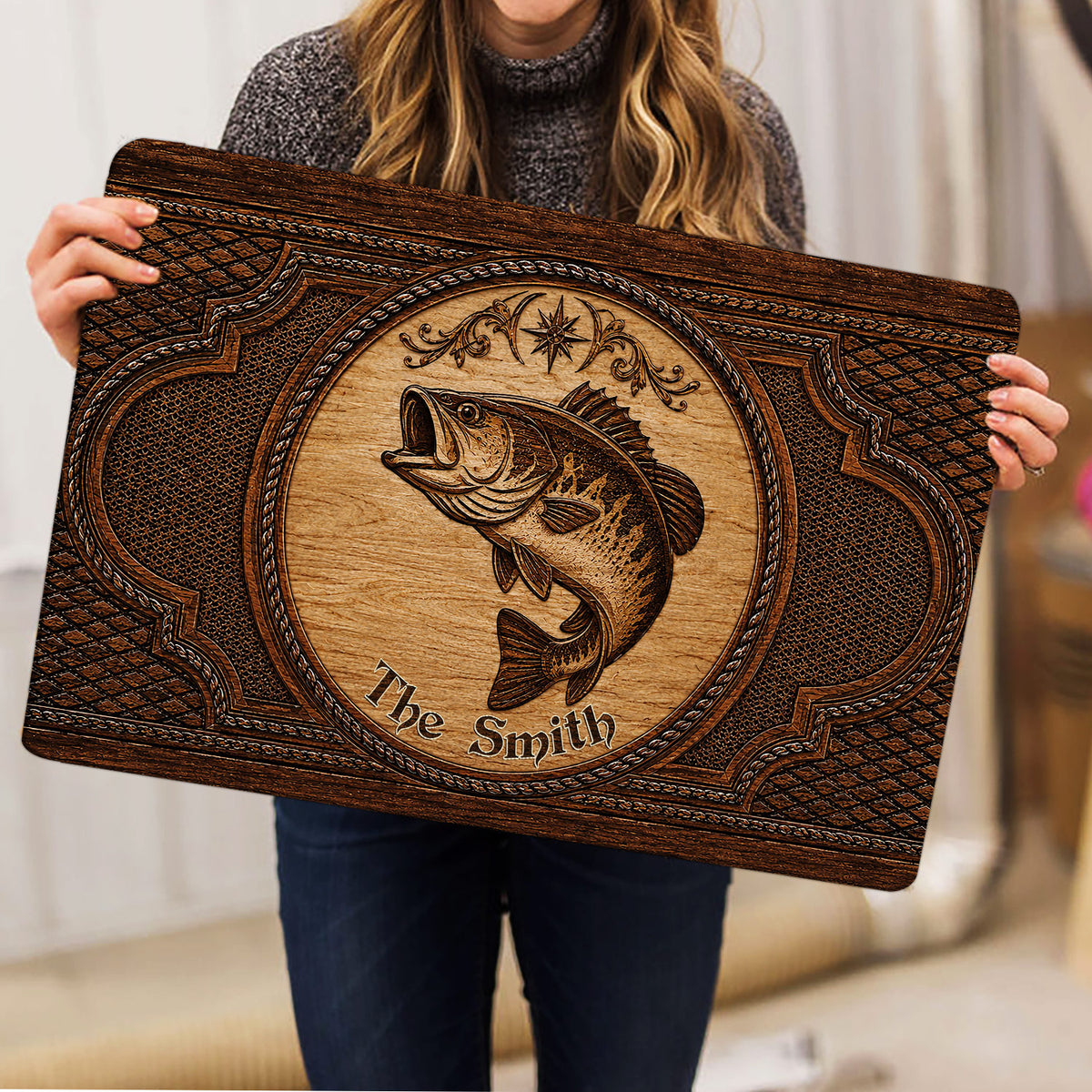 Person holding a rustic fishing doormat featuring a central fish illustration and ornate brown border
