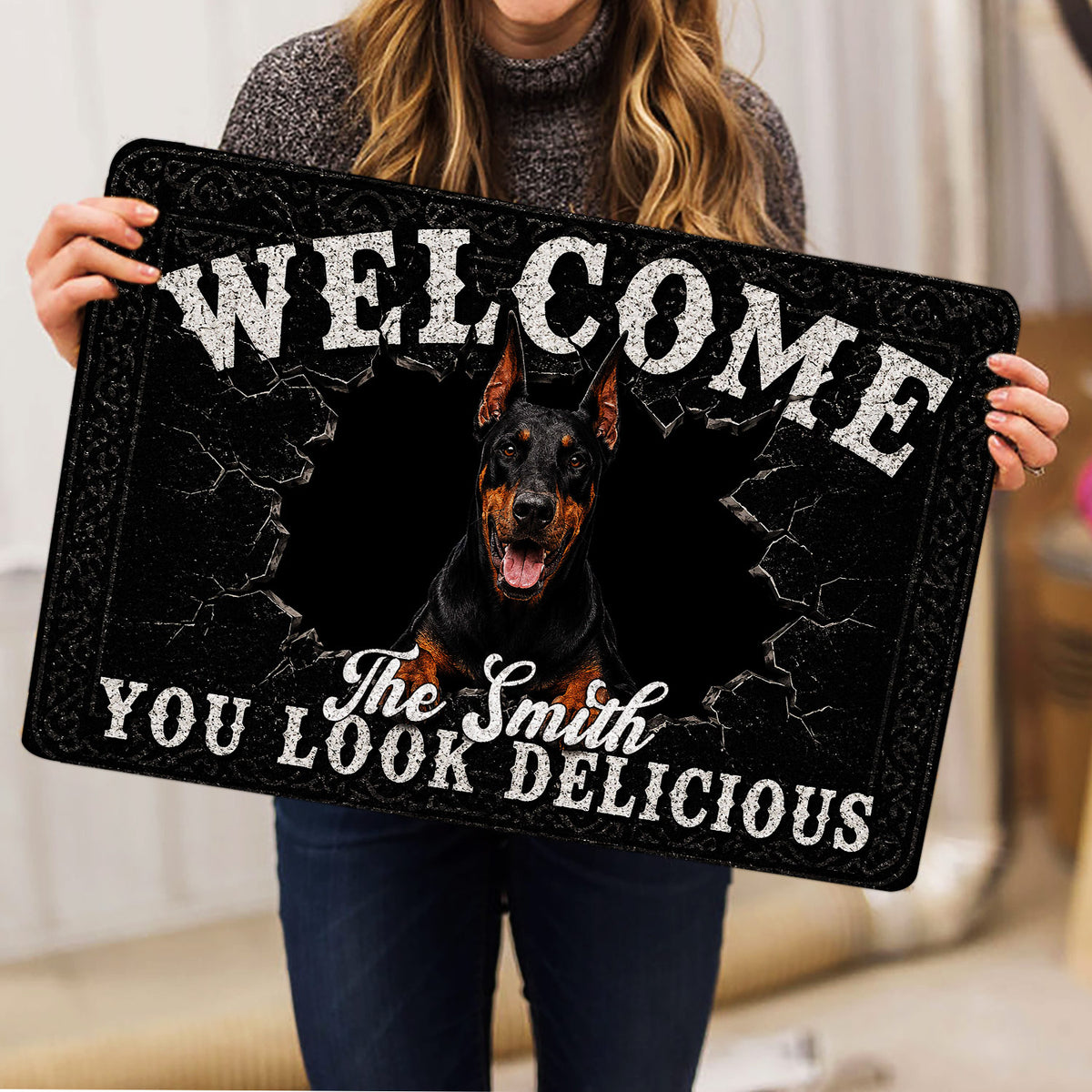 Person holding a black Doberman doormat featuring cracked stone artwork and bold welcome lettering