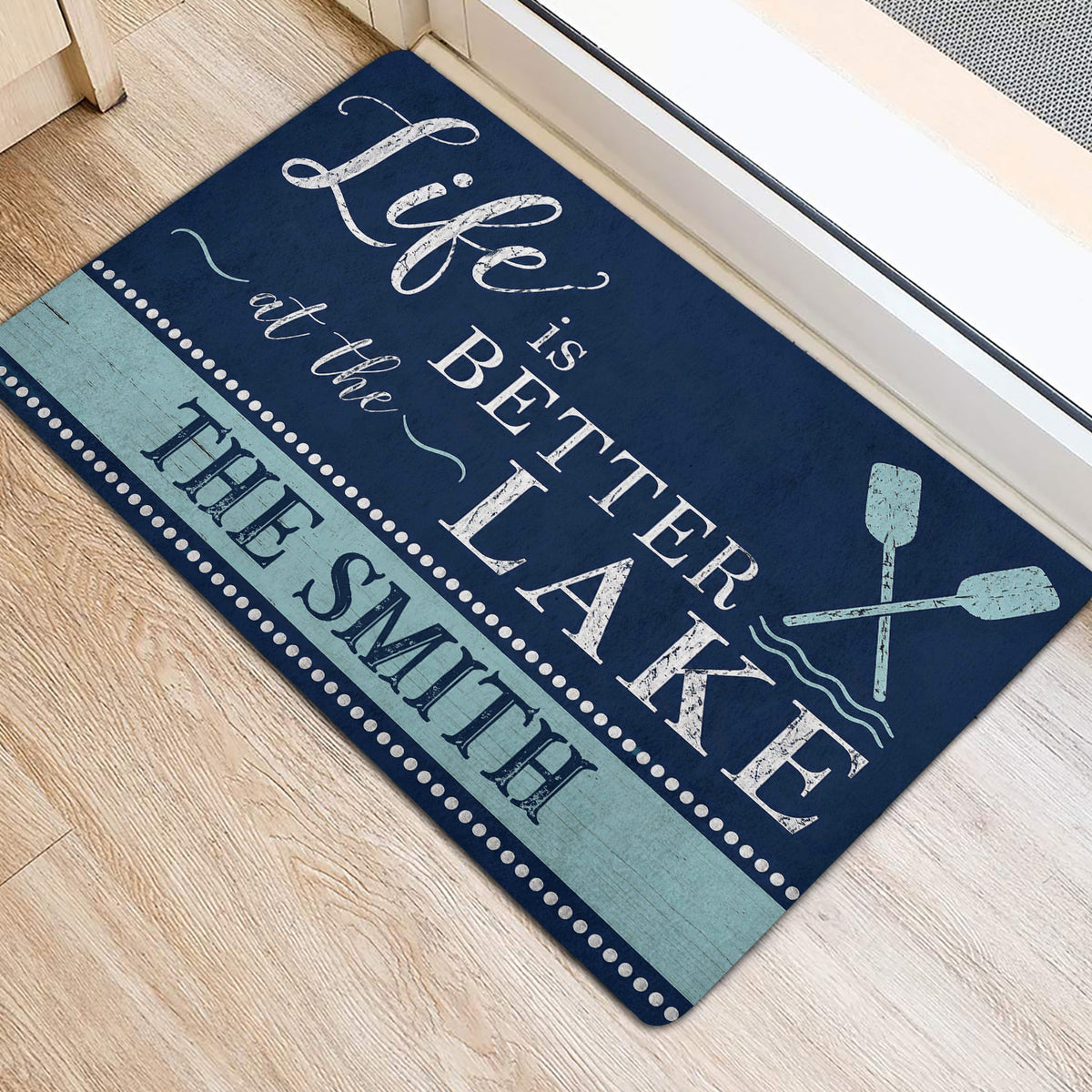 Lake-themed entrance mat positioned beside a doorway, featuring navy lettering, aqua accents and oars