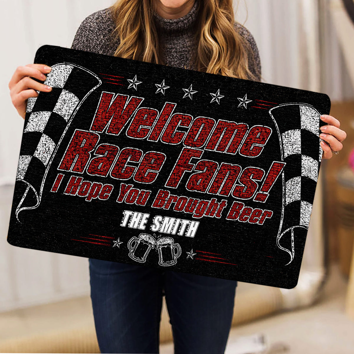 Person holding a black racing doormat featuring bold red lettering, checkered flags, and beer mugs