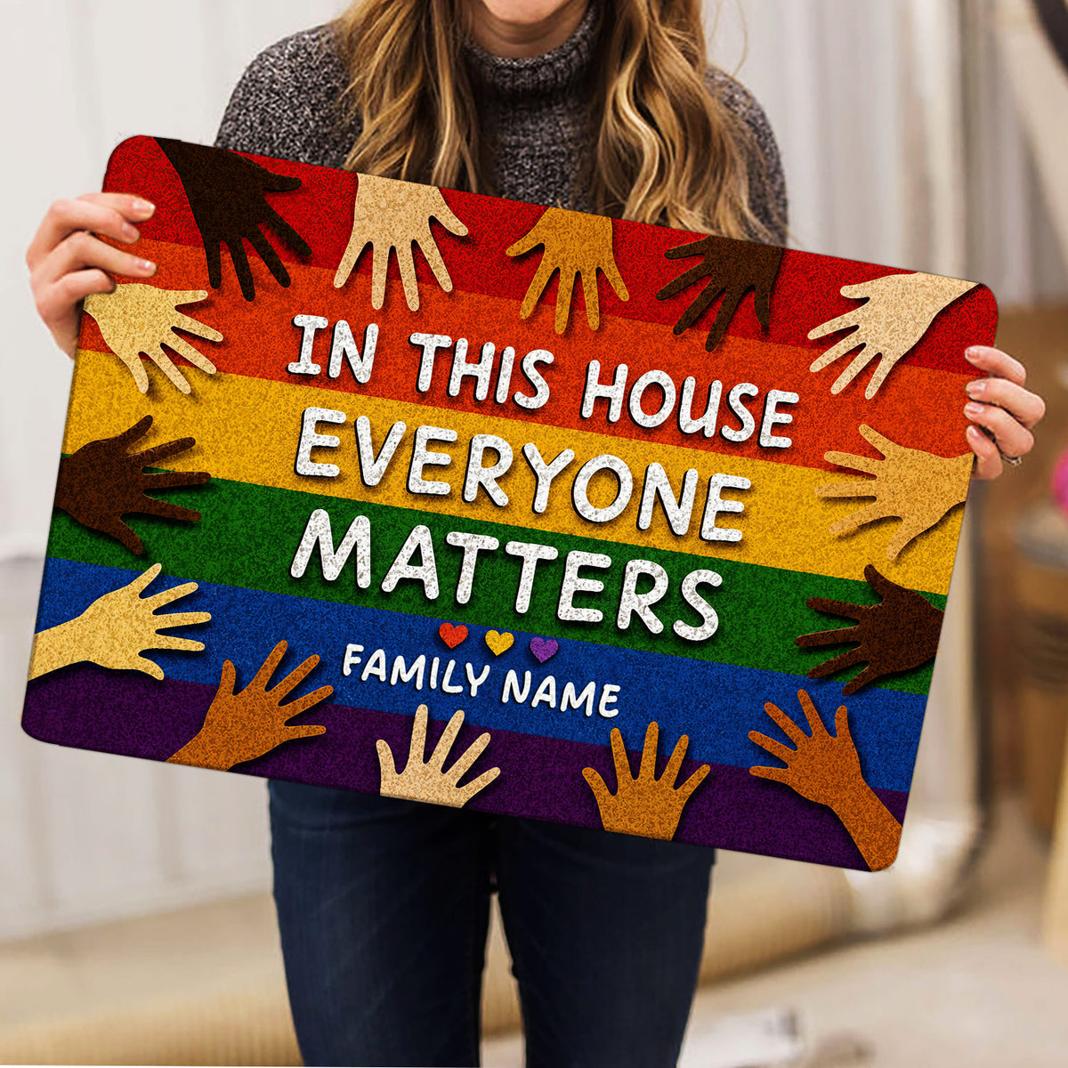 Person holding a colorful LGBT doormat featuring rainbow stripes, diverse hands, and an inclusive message