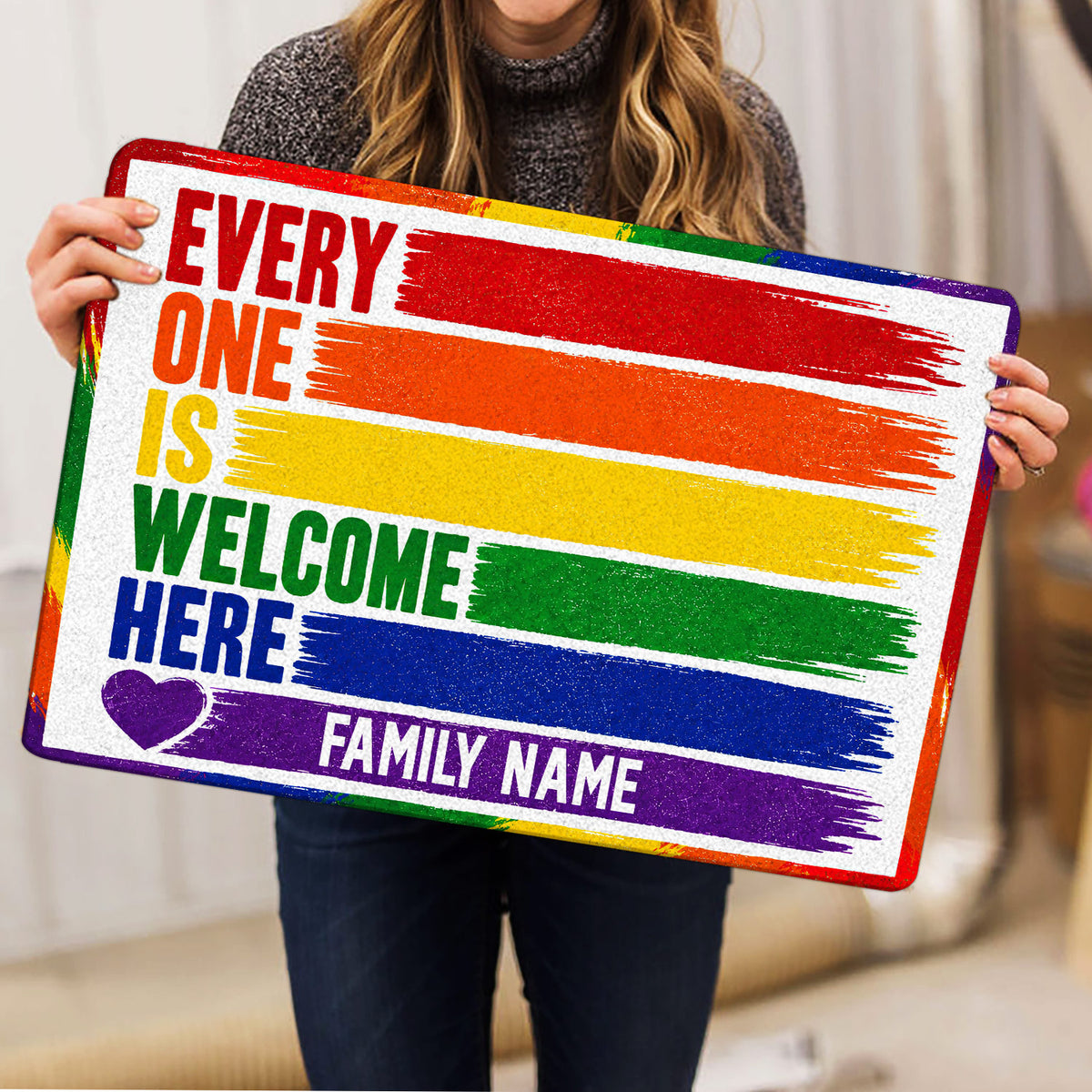 Person holding a colorful LGBT pride doormat featuring rainbow brushstrokes, welcome wording, and a heart