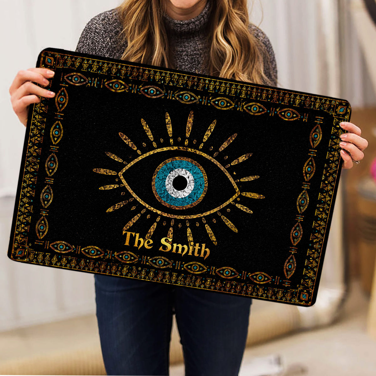 Person holding a black evil eye doormat featuring blue, white, and gold decorative artwork