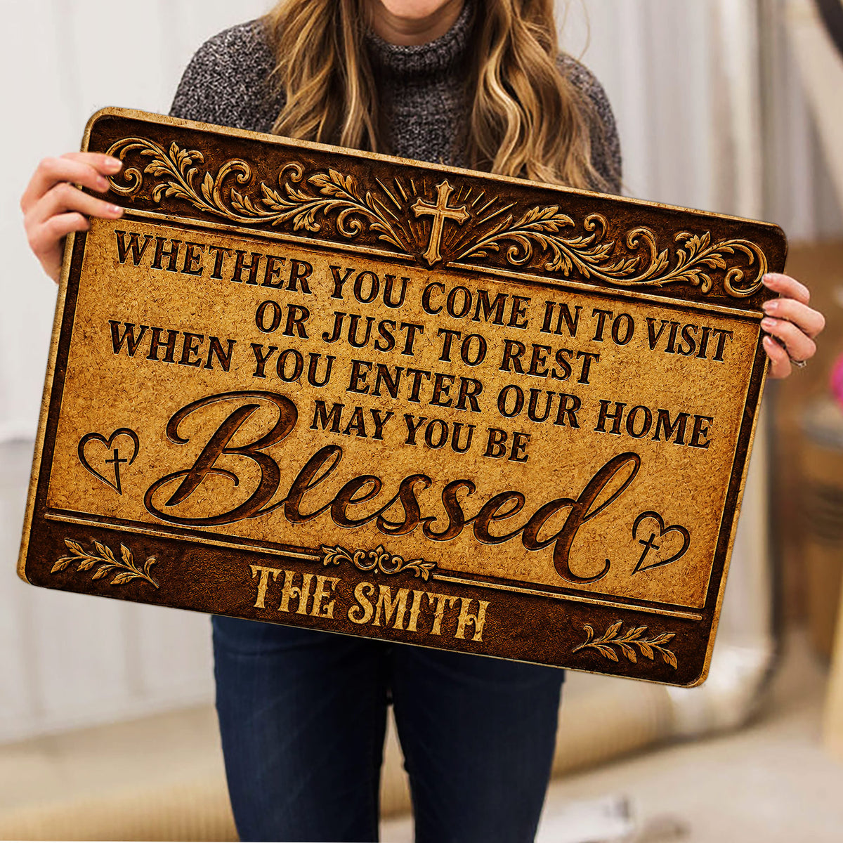 Woman holding an ornate Christian doormat featuring a cross, hearts, and a welcoming message