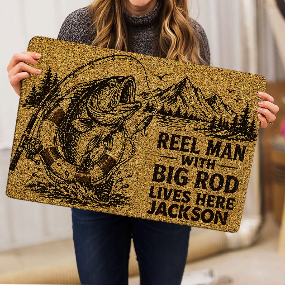 Person holding a fishing doormat featuring a large fish, rod, lure, mountains, and rustic brown design