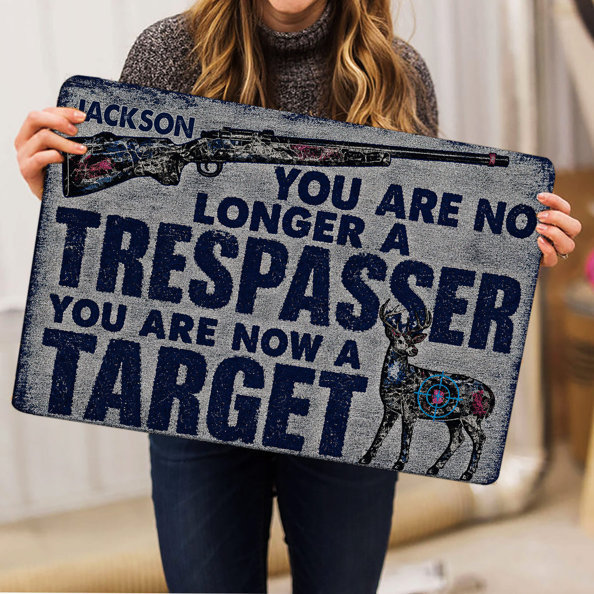 Person holding a hunting doormat featuring a rifle, deer illustration, and bold distressed lettering
