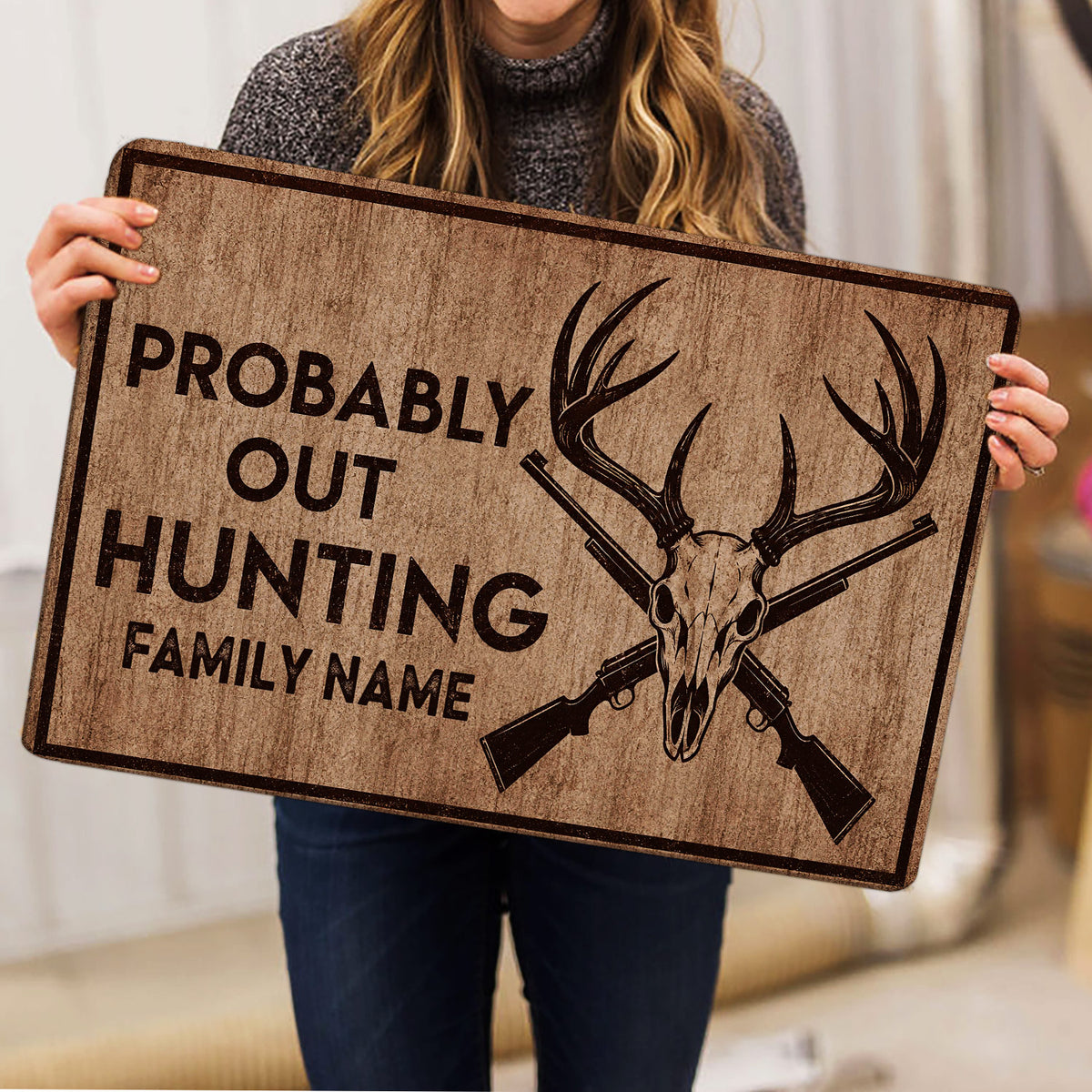 Person holding a rustic hunting doormat featuring antlers, deer skull, crossed rifles, and bold lettering