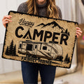 Person holding a rustic camping doormat featuring a travel trailer, mountains, trees, and bold lettering
