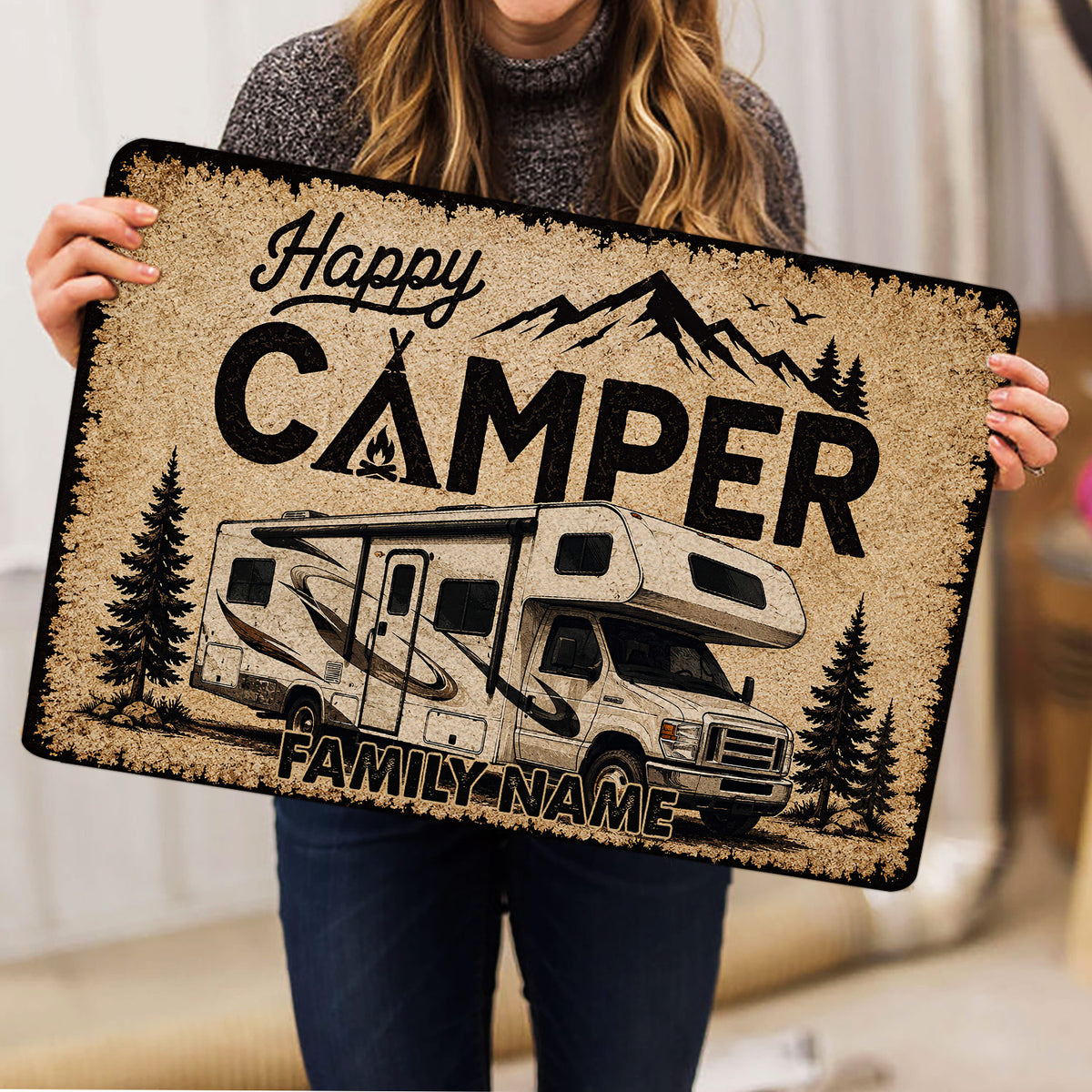 Person holding a camping doormat with an RV, mountains, pine trees, and family welcome artwork