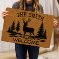 Person holding a tan deer hunting doormat featuring pine trees and welcome artwork