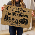 Person holding a camping doormat featuring mountains, pine trees, camp chairs, campfire, and travel trailer artwork