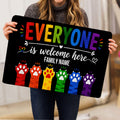 Person holding a black LGBT doormat featuring rainbow lettering, a welcome message, and colorful paw prints