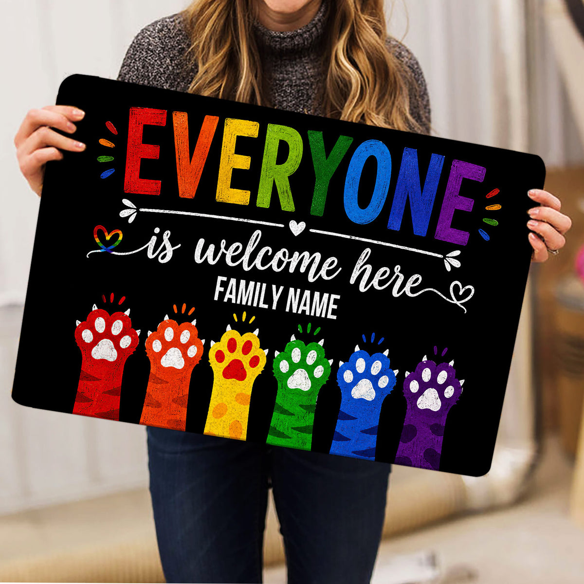 Person holding a black LGBT doormat featuring rainbow lettering, a welcome message, and colorful paw prints