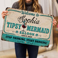 Person holding a vintage-style mermaid saloon doormat featuring turquoise lettering and cocktail artwork