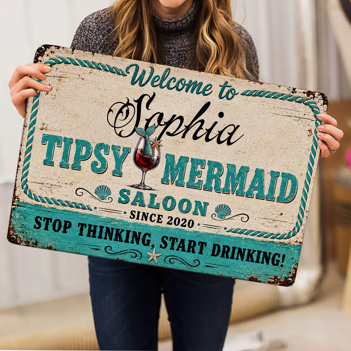 Person holding a vintage-style mermaid saloon doormat featuring turquoise lettering and cocktail artwork