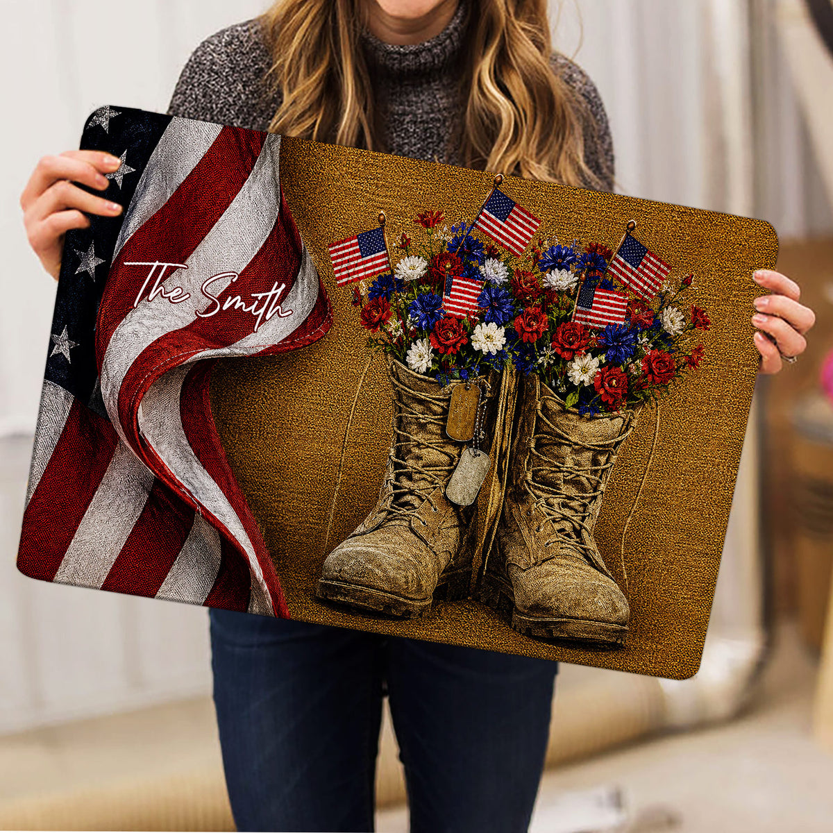 Person holding a patriotic veteran doormat featuring combat boots, flags, flowers, and a folded American flag