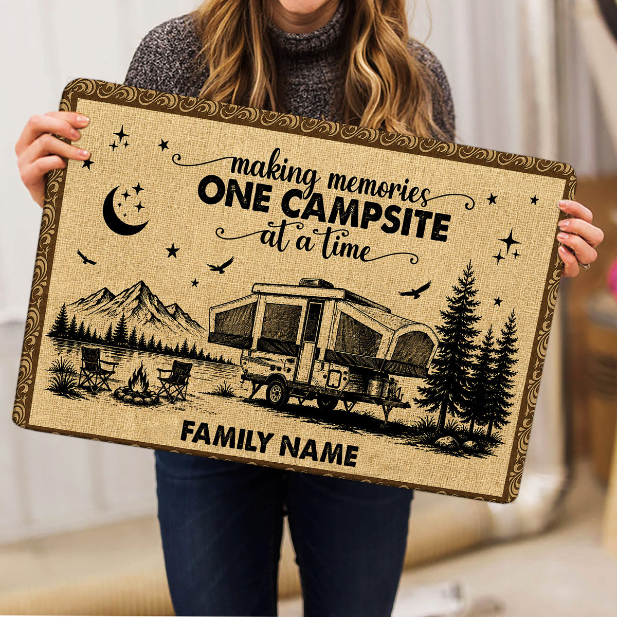Person holding a rustic camping doormat featuring an RV campsite, mountains, trees, stars, and campfire design