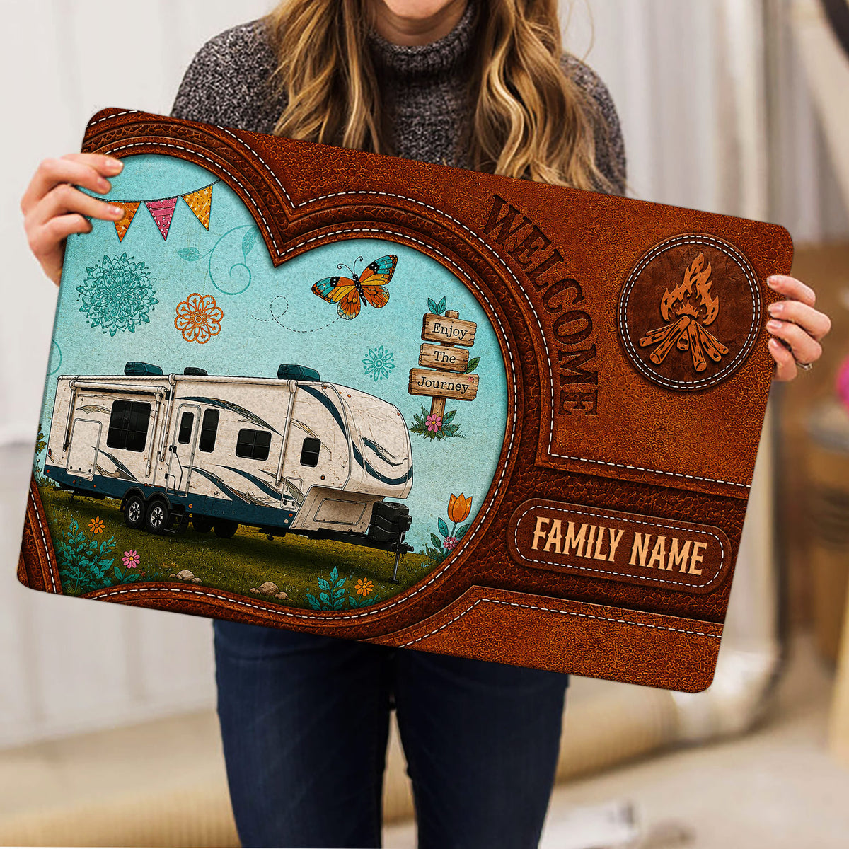 Camping-themed doormat held upright, showing a travel trailer, campfire emblem, floral details, and rustic border