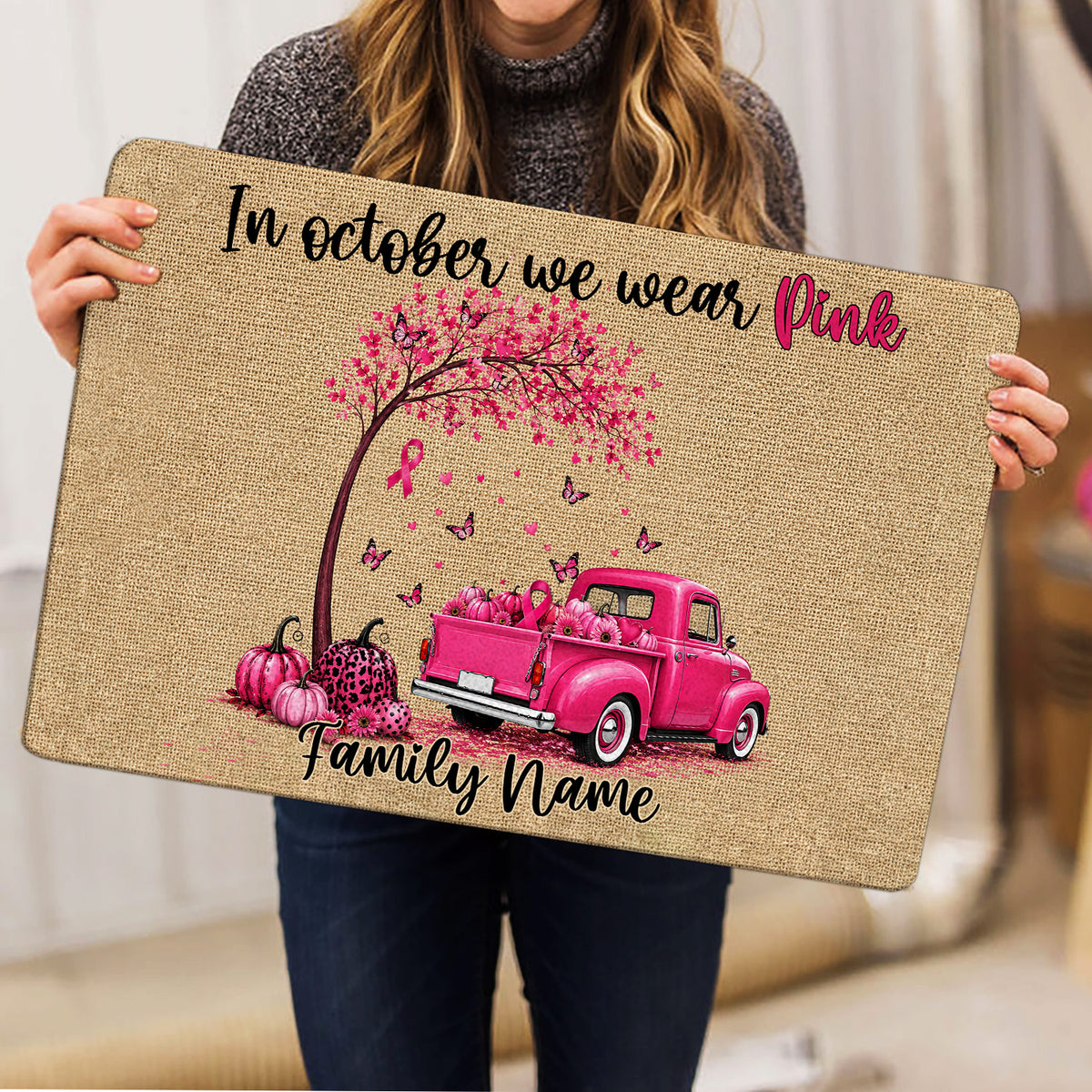 Woman holding a breast cancer awareness doormat featuring a pink ribbon tree, pumpkins, and pink truck