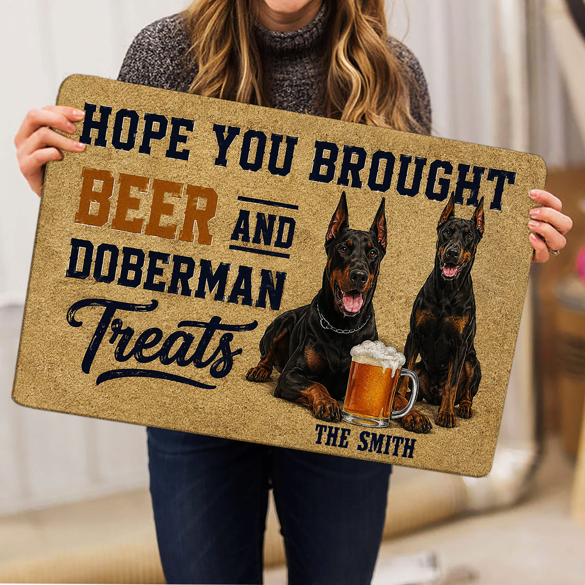 Person holding a Doberman doormat featuring two dogs and a beer-themed design