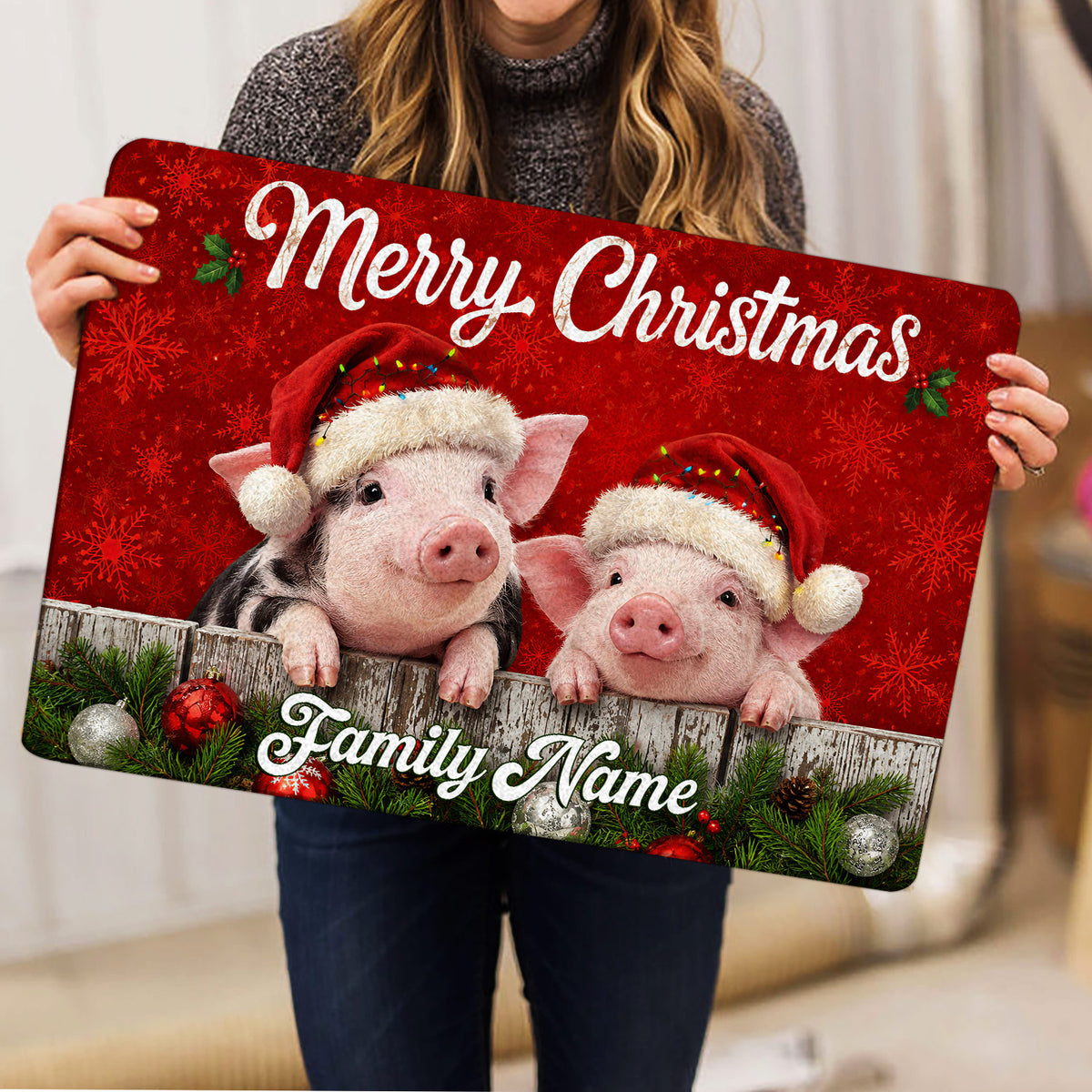 Two pigs wearing Santa hats featured on a red Christmas doormat held indoors with holiday greenery and ornaments