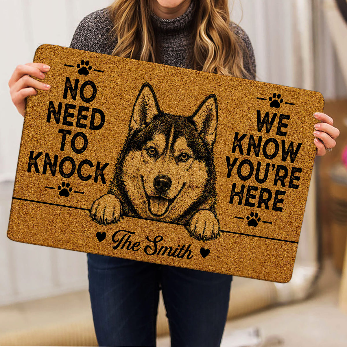 Person holding a tan husky doormat featuring paw prints and a humorous greeting