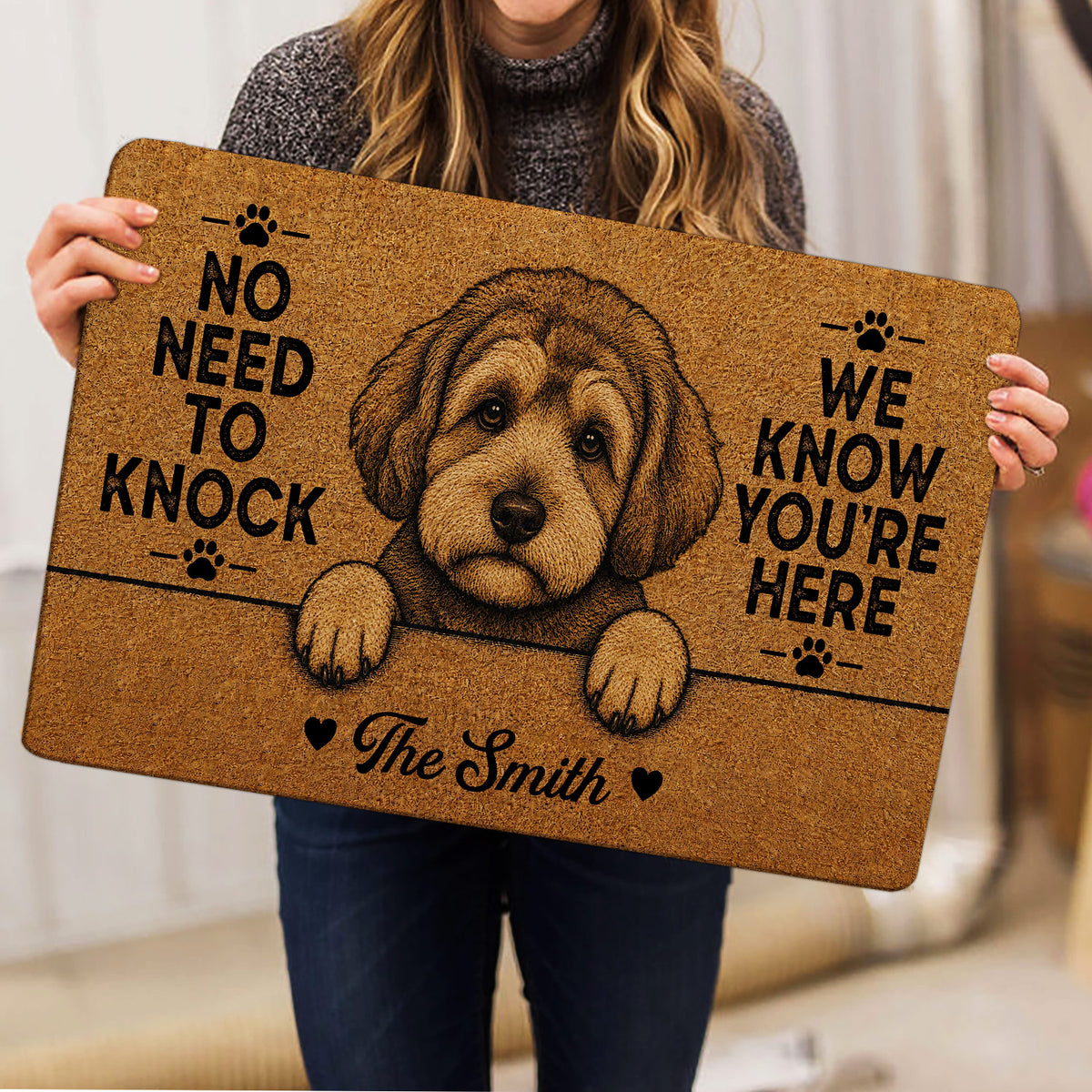 Person holding a tan Schnoodle doormat with dog artwork, paw prints and bold black lettering