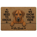 Custom family name Rhodesian Ridgeback dog doormat featuring a reddish brown floppy-eared dog peeking over a line, black paw prints, natural coir-look background and No Need to Knock We Know You’re Here quote.