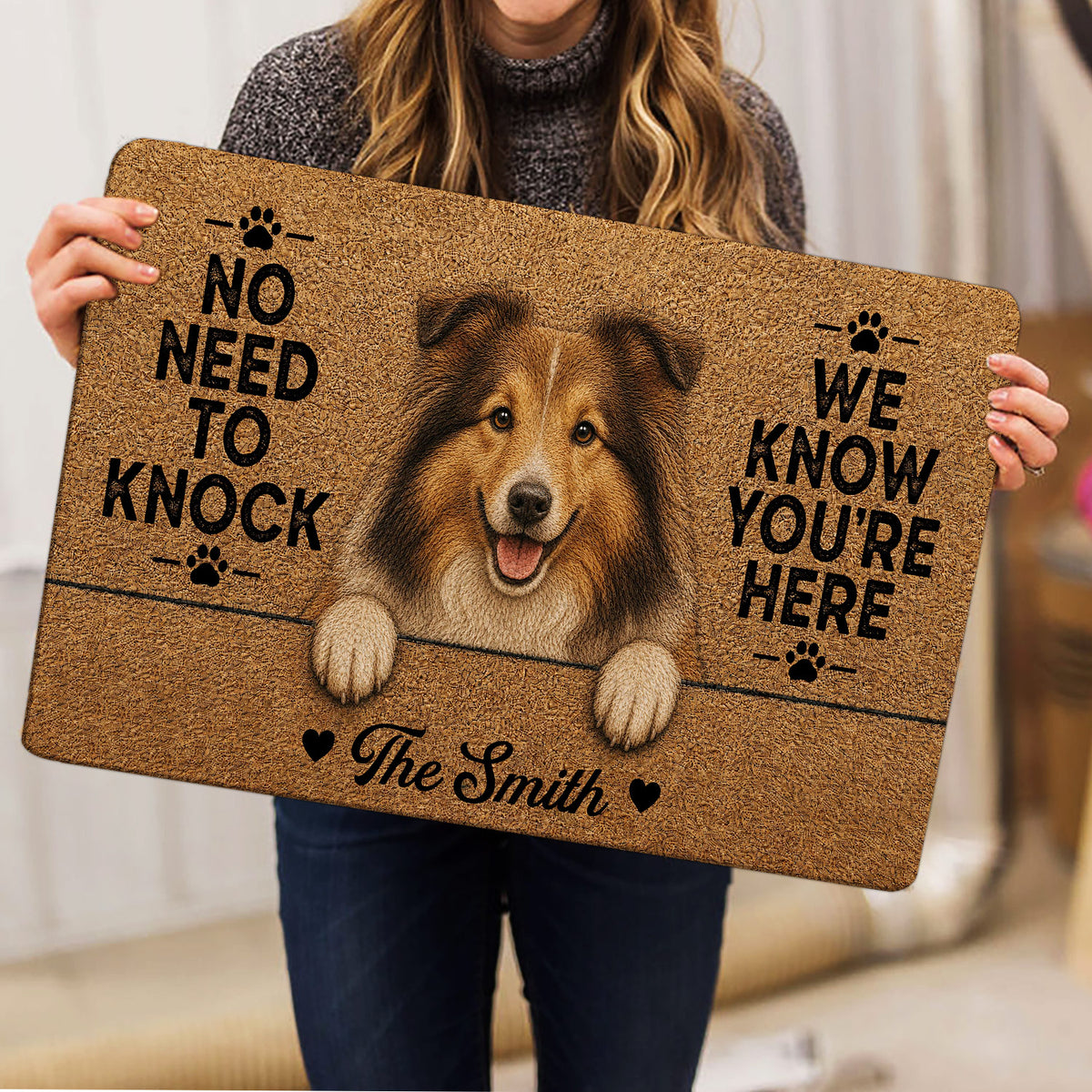 Person holding a Sheltie doormat showing a smiling dog portrait, paw prints, warm brown artwork, and entryway lettering