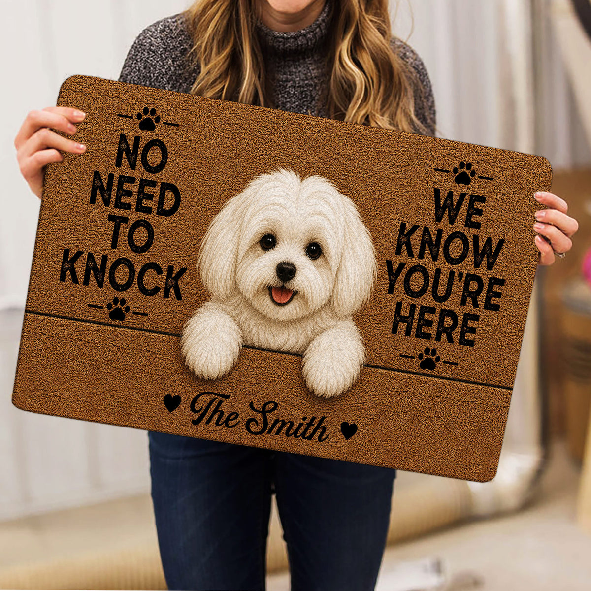 Person holding a Maltese dog doormat featuring a white dog portrait, paw prints, hearts, and warm brown artwork
