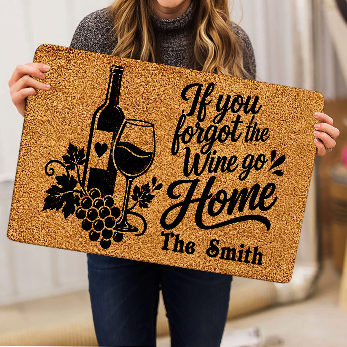 Person holding a wine-themed doormat featuring a bottle, wine glass, grapes, and script artwork