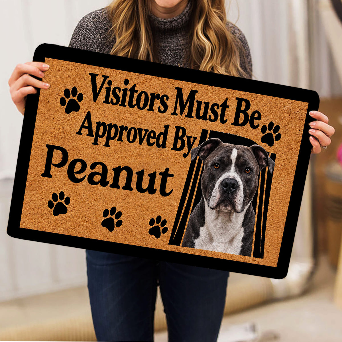 Person holding an American Staffordshire dog doormat with a tan background and black paw print accents