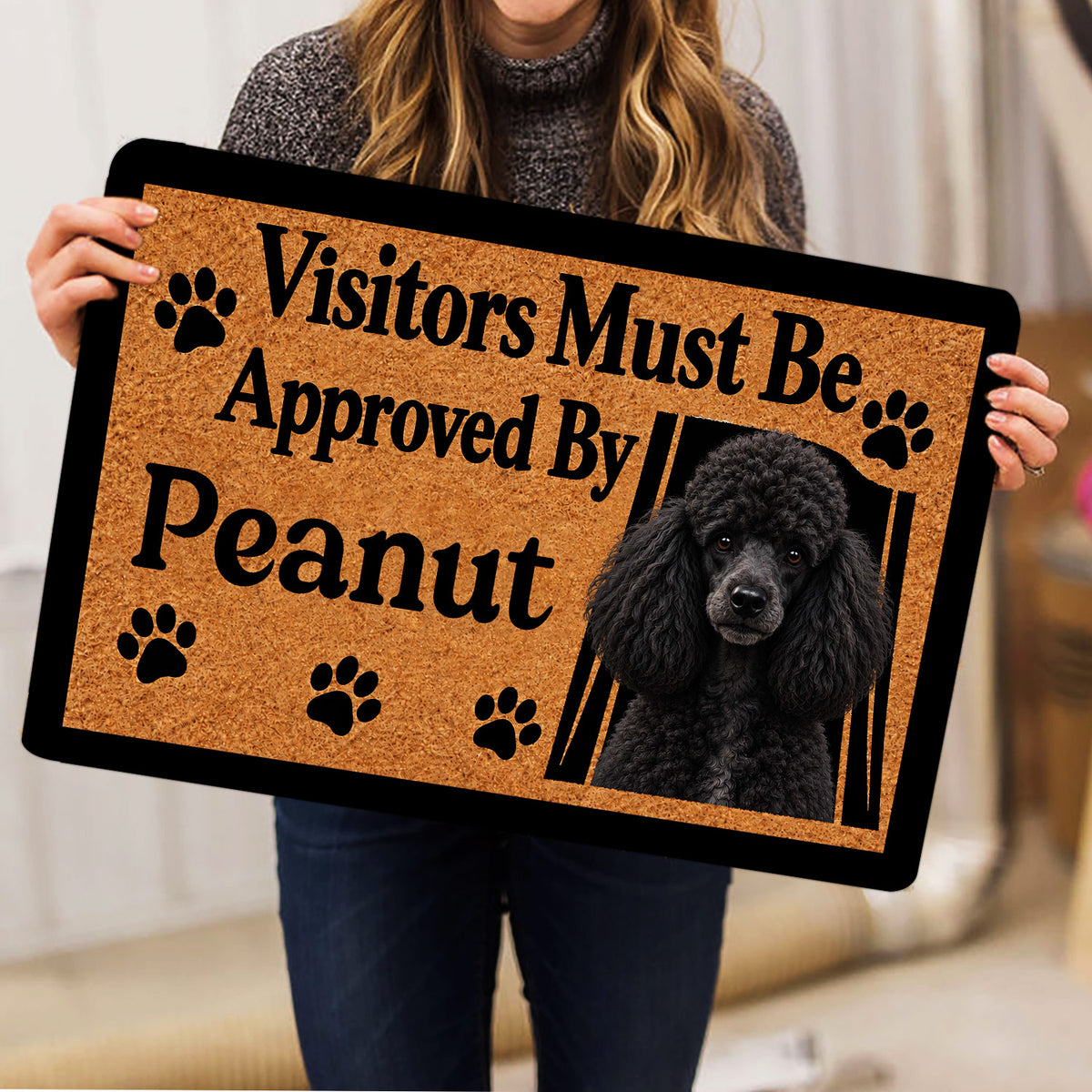 Person holding a poodle doormat featuring a black curly dog portrait, paw prints, and tan artwork