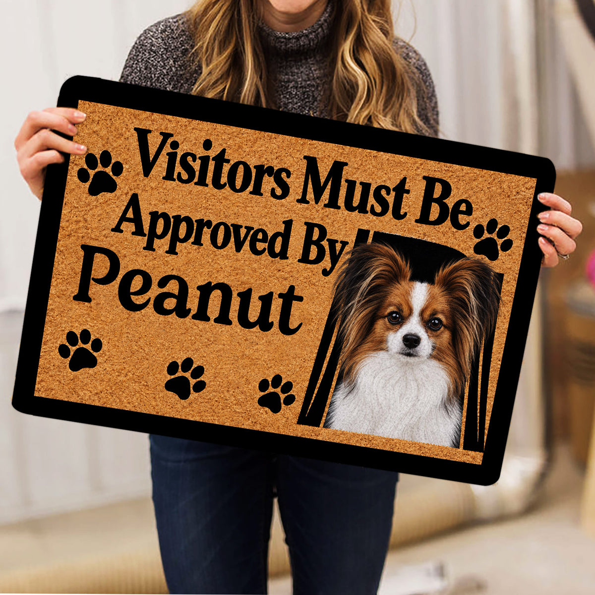 Person holding a Papillon dog doormat featuring a portrait, paw prints, and bold black lettering