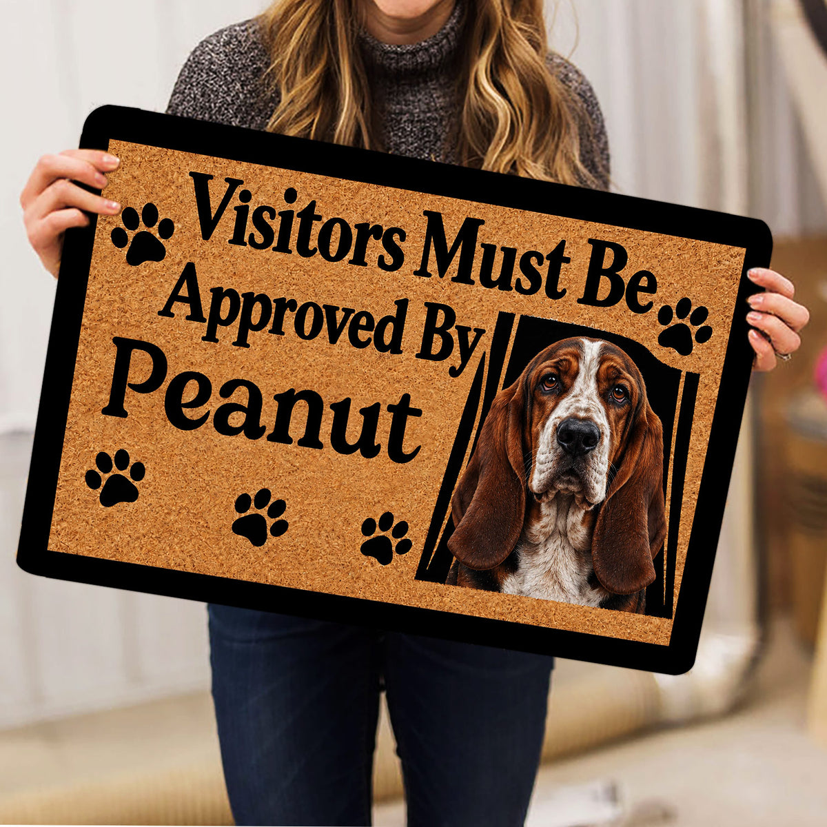 Person holding a Basset Hound doormat featuring paw prints and a friendly visitor message