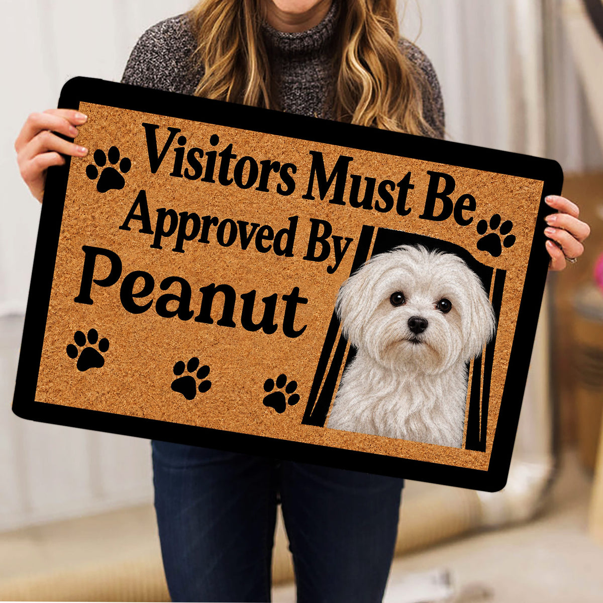 Maltese dog doormat held indoors, featuring a white dog portrait, paw prints, and bold black lettering