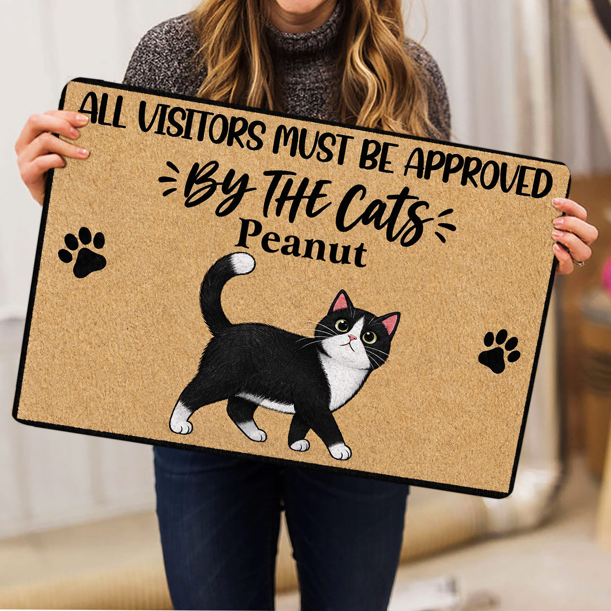 Person holding a tuxedo cat doormat featuring paw prints and a playful cat-themed message