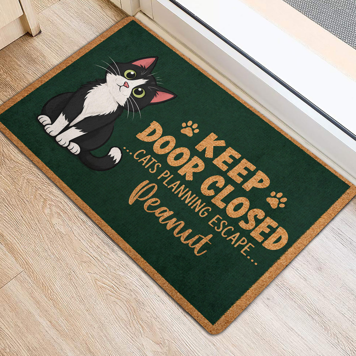 Tuxedo cat door mat positioned near a doorway, featuring dark green artwork and tan decorative edging