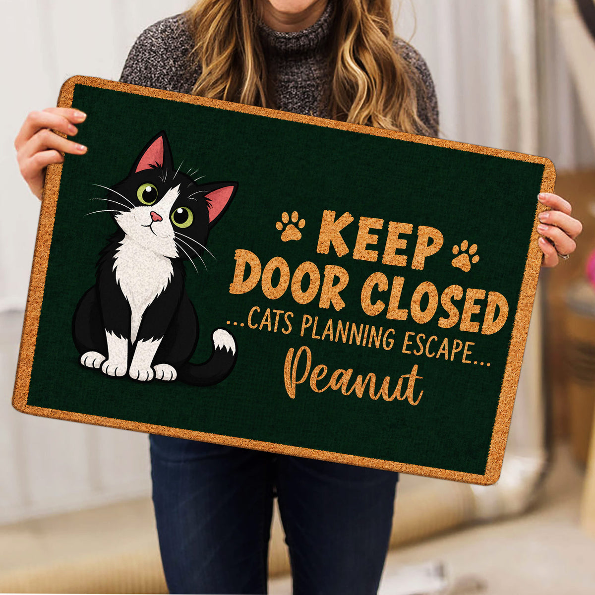 Tuxedo cat doormat held indoors, showing the full dark green design and humorous cat-themed artwork