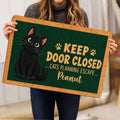 Person holding a black cat doormat featuring paw prints, green artwork and a playful entryway message