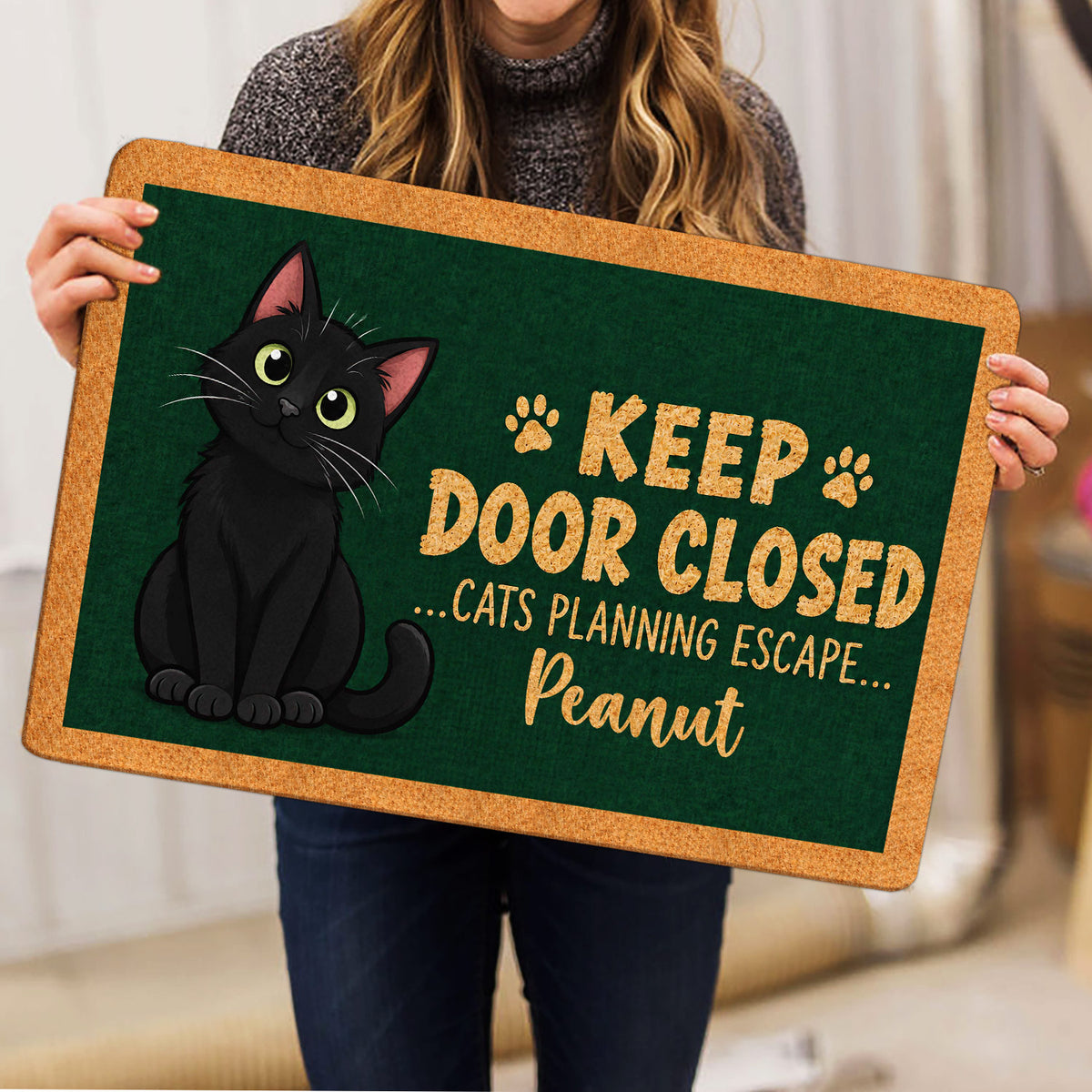 Person holding a black cat doormat featuring paw prints, green artwork and a playful entryway message