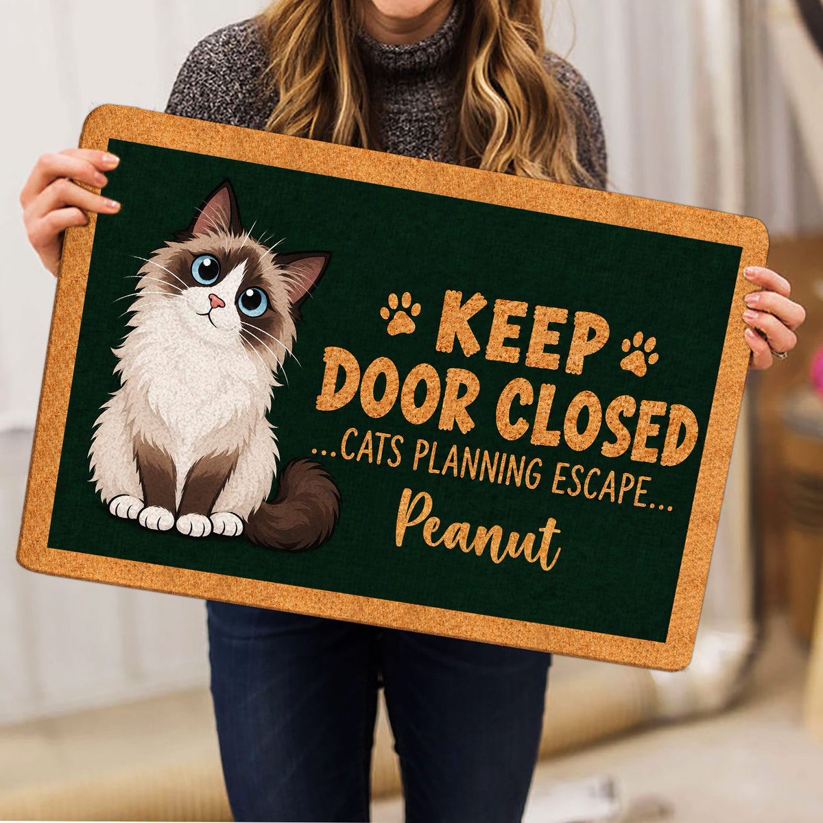 Person holding a Ragdoll cat doormat featuring a fluffy blue-eyed cat and paw print artwork