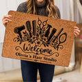 Person holding a tan hairstylist doormat featuring black hair tools, flowers, and welcome artwork