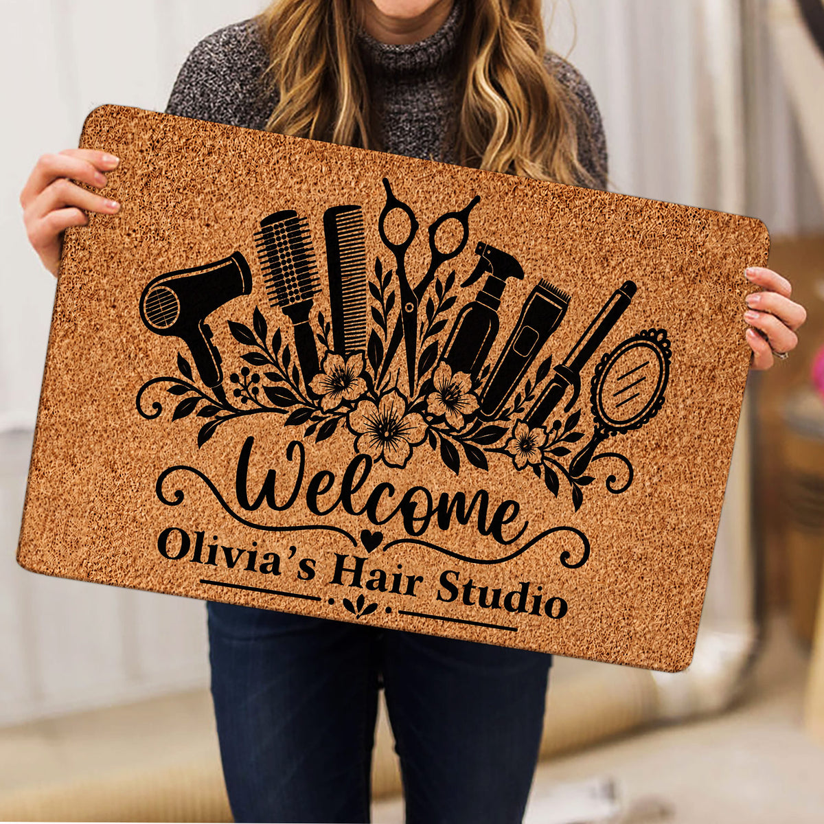 Person holding a tan hairstylist doormat featuring black hair tools, flowers, and welcome artwork
