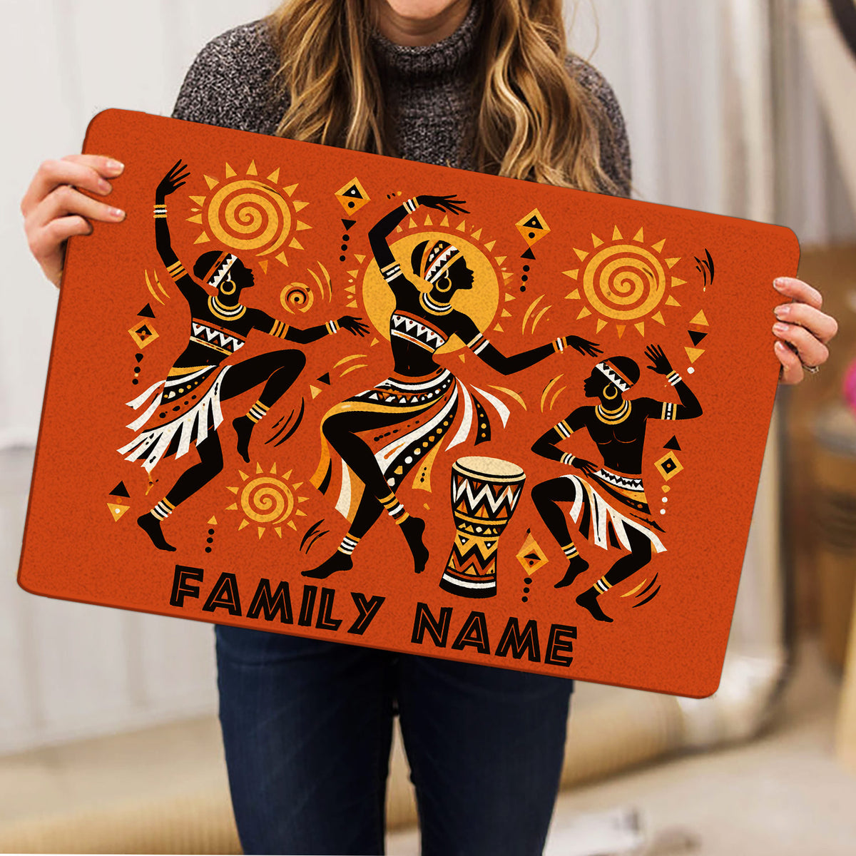 Person holding an orange African-inspired doormat featuring dancers, sun symbols, geometric details, and a drum