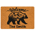 Bear silhouette doormat with mountain, pine tree, moon, and welcome artwork in black on a natural brown background