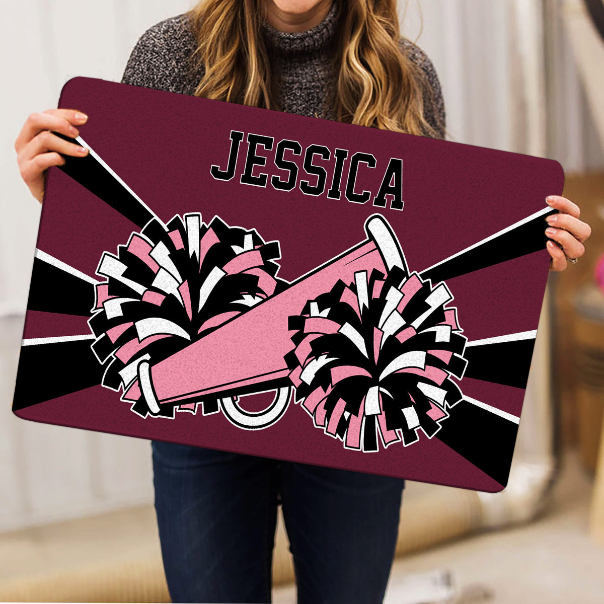 Person holding a cheerleading doormat with colorful pom-poms and megaphone artwork in a home setting