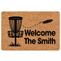 Disc golf doormat with a basket, flying disc, and bold welcome message in black on a tan background