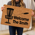 Person holding a disc golf doormat featuring a basket, flying disc, and bold welcome artwork